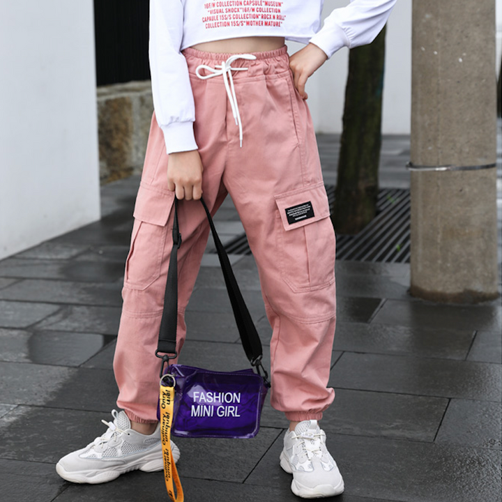 Une personne porte un pantalon rose et une chemise blanche. Elle porte également un sac à main violet et tient une pancarte rectangulaire noire avec du texte blanc. On peut voir de près une chaussure et une bande métallique.