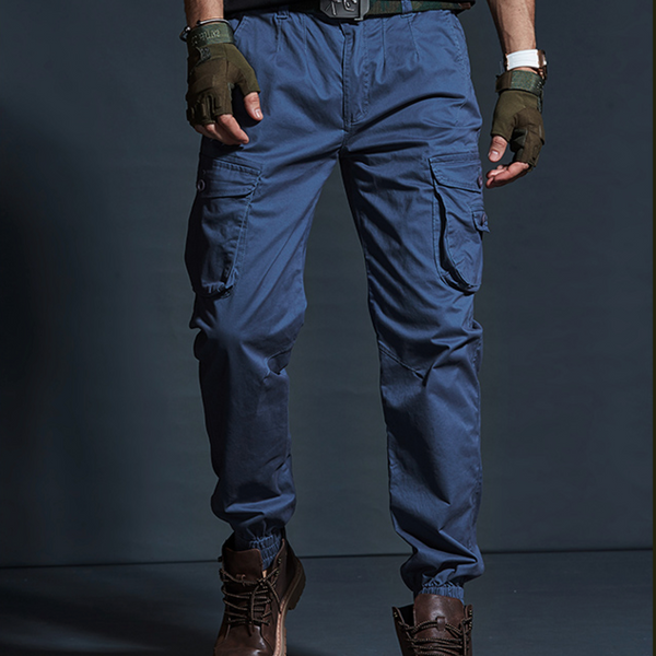 Un homme porte un pantalon cargo technique bleu, des boots marrons, et des mitaines kaki.