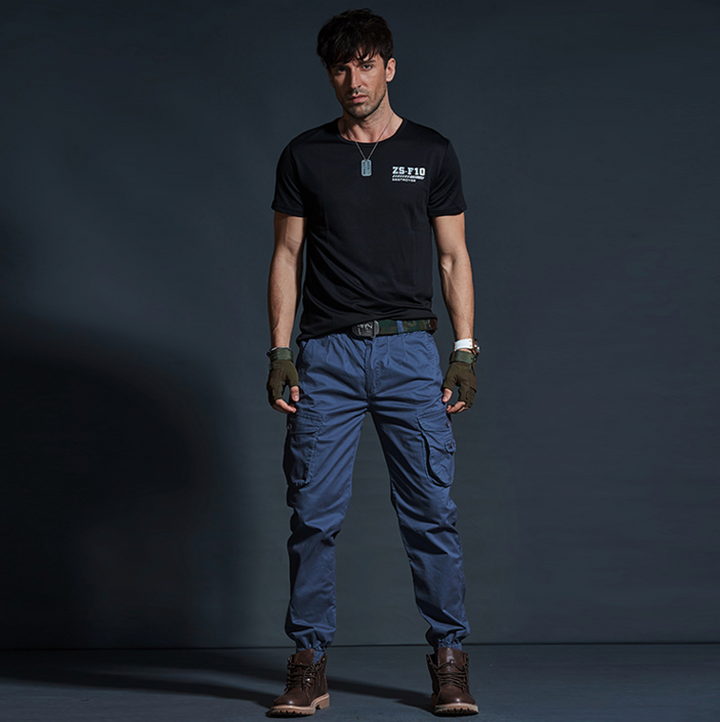 Un homme porte un pantalon cargo en coton bleu avec des chevilles resserrées. Le pantalon est de style militaire avec des poches cargo latérales. Disponible en tailles 28 à 40.