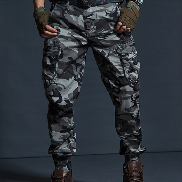 Un homme porte un pantalon cargo technique gris camo, des boots marrons  et des mitaines kakis.