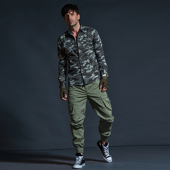 Un homme porte un pantalon cargo en coton kaki avec des chevilles resserrées. Le pantalon est de style militaire avec des poches cargo latérales. Disponible en tailles 28 à 40.