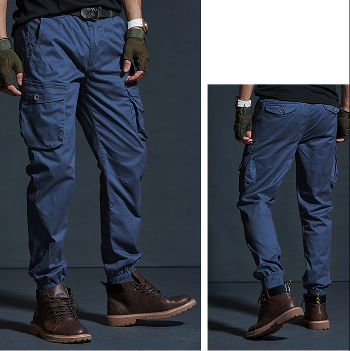 Un homme porte un pantalon cargo bleu avec des bottes marron. Le pantalon a des poches cargo latérales et des chevilles resserrées. Pantalon cargo coton resserré aux chevilles - Bleu - Homme.