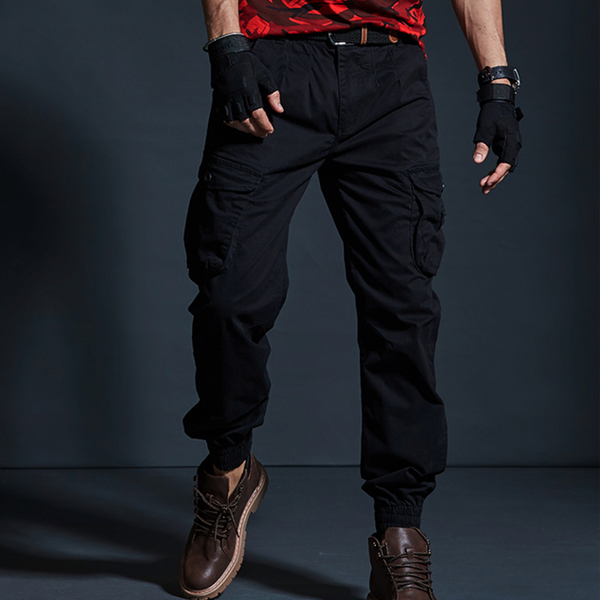 Un homme porte un pantalon cargo technique noir, des boots marrons, un tee-shirt rouge et des mitaines noires.