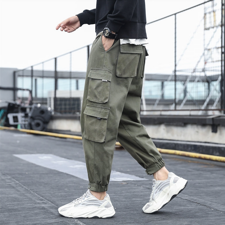 Un homme porte un pantalon cargo kaki ample avec des poches multiples et resserré aux chevilles. Le pantalon est en coton 100% pour un confort maximal et une durabilité garantie. Disponible en tailles S à 5XL.