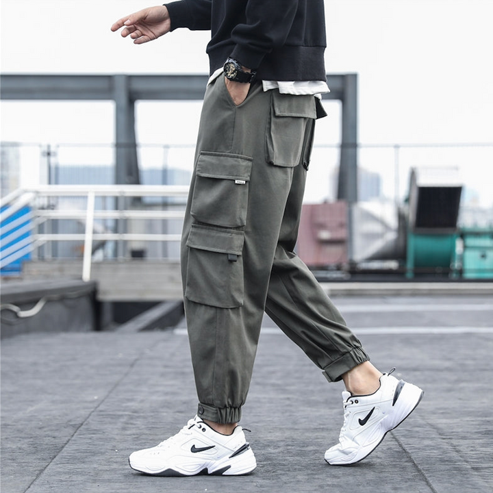 Un homme porte un pantalon cargo ample multipoches resserré aux chevilles - Gris. Coupe tendance et pratique avec élastique à la taille et cordon ajustable. Fabriqué en coton 100% pour confort et durabilité. Tailles disponibles du S au 5XL.