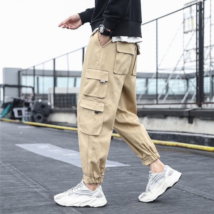 Un homme porte un pantalon cargo ample multipoches resserré aux chevilles beige de la marque Cargo District. Ce pantalon en coton 100% offre confort et durabilité, avec une coupe ajustable du S au 5XL.