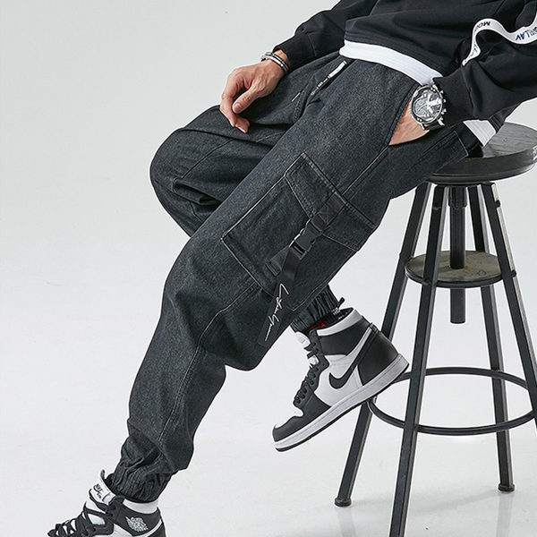 Un homme pose sur un tabouret noir avec un sweat noir, des baskets blanches et noires et un jean cargo resserré aux chevilles avec des poches à fermeture clip. Le jean est noir.