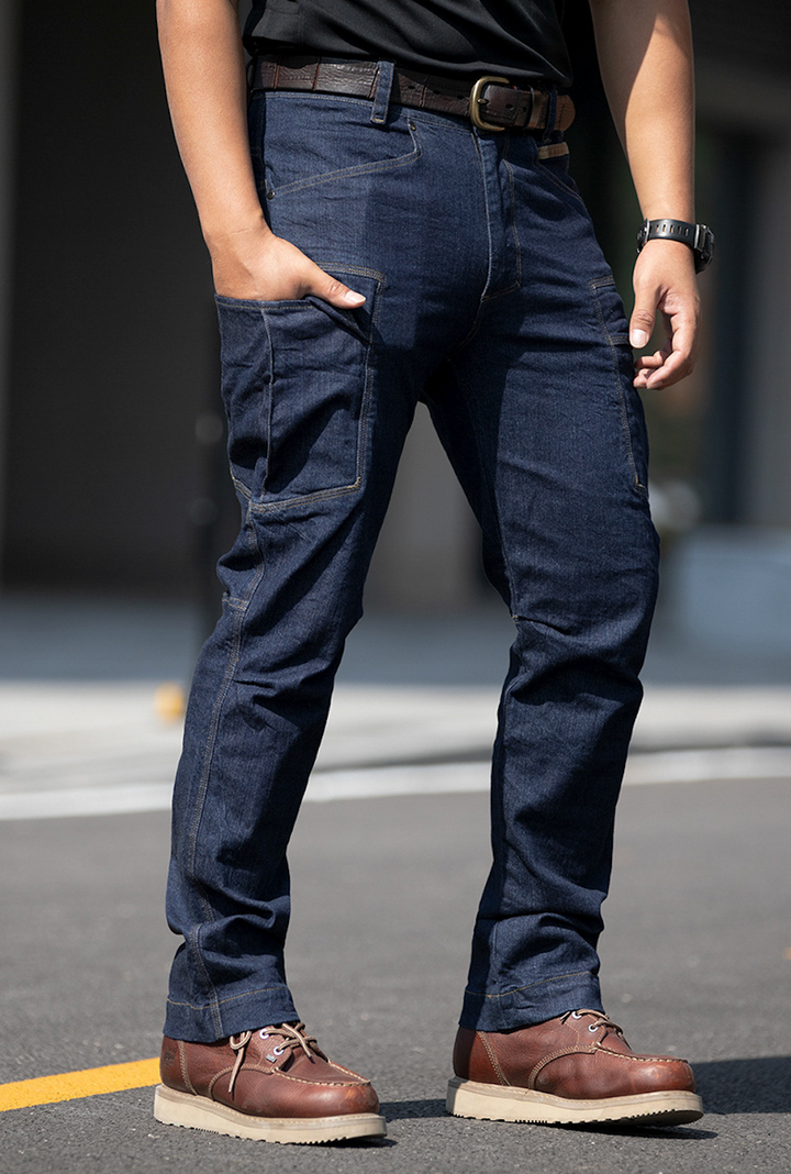 Un homme portant un jean cargo ajusté coupe droite - Bleu. Le jean en toile denim stretch offre un confort optimal et une grande liberté de mouvement. Nombreuses poches pour une fonctionnalité pratique. Subtile touche d'originalité avec une couture camo kaki sur l'une des poches à la taille. Tailles disponibles du S au 2XL.