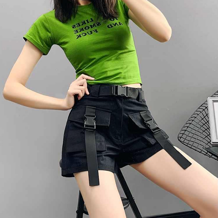 Une femme pose derrière un mur gris. Elle pose sur un tabouret noir. Elle porte un tee-shirt vert et un short cargo taille haute noir avec ceinture noire et lanières noires à clip sur les poches.