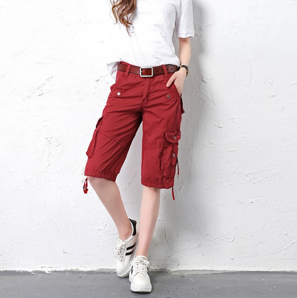 Une femme pose devant un mur extérieur blanc. Elle porte un tee-shirt blanc, des baskets blanches et un short cargo long, coupe droite rouge bordeaux. 