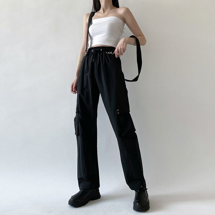 Une femme porte un pantalon cargo noir avec des sangles bretelles amovibles. Le pantalon est taille haute avec une coupe droite et une taille élastique pour un ajustement confortable. Disponible du S au 3XL. Le pantalon ajoute une note d'originalité à votre style urbain et street.