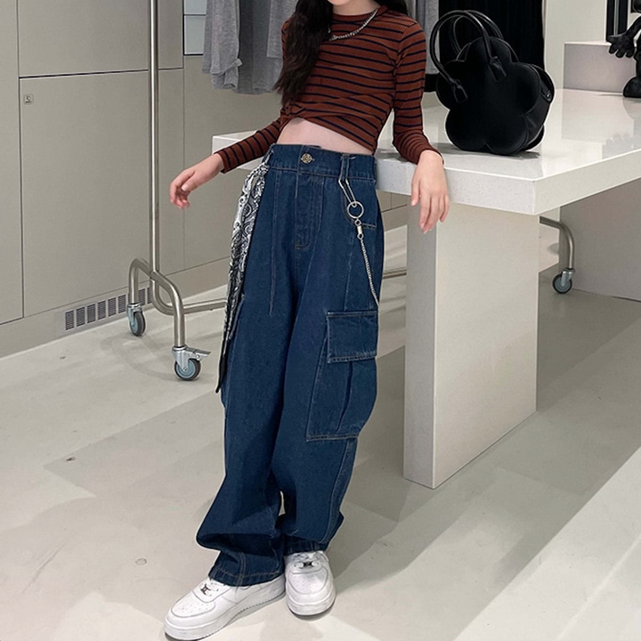 Une fille pose pour une photo, portant un pantalon cargo jean foulard chaîne bleu. Le pantalon a une coupe ample avec des poches latérales pour plus de commodité. Disponible du 4 au 14 ans.