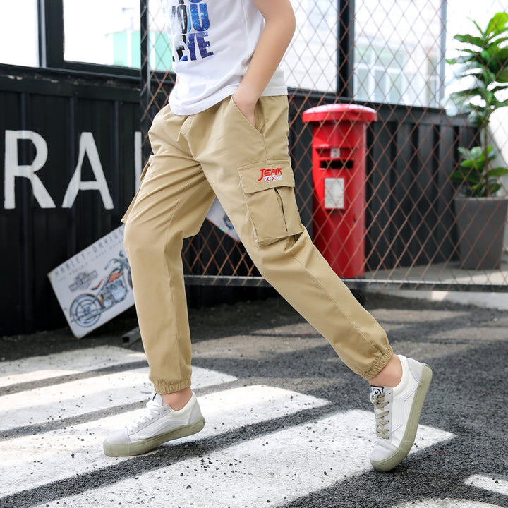 Un garçon pose dans un hangar. Il y a une plante derrière lui et un poster de moto. Il porte un tee-shirt blanc avec imprimé bleu, des baskets blanches ainsi qu'un pantalon cargo jogger beige.