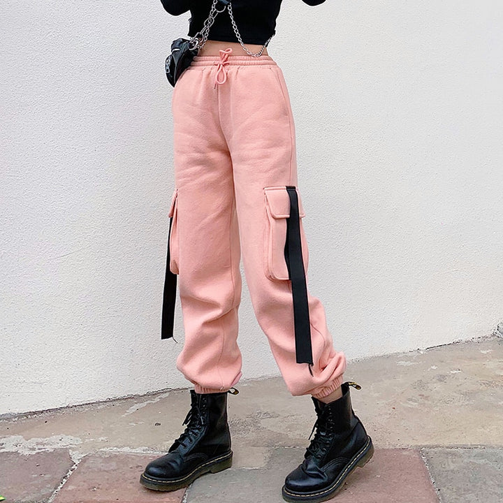 Une femme porte un Pantalon cargo jogger rose avec des lanières noires sur les poches latérales. Coupe confortable et taille haute pour un look urbain et féminin. Disponible du S au L.