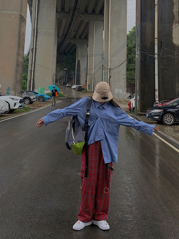 Une personne portant un chapeau et une chemise bleue se tient sur une route avec un pont au-dessus. Elle porte un Pantalon cargo à carreaux et chaînes rouge, avec une coupe évasée et une jambe large. Le pantalon est orné d'une chaîne métallique, d’œillets en métal et de lanières en similicuir, ainsi que d'épingles sur les côtés pour une touche décalée. Disponible du S au L.