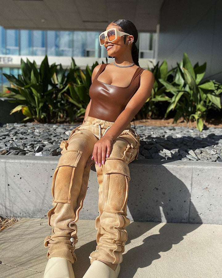 Une femme pose dans la rue devant des plantes. Elle porte des lunettes de soleil, des chaussures beige, un top en similicuir marron et un pantalon cargo jogger tie and dye beige.