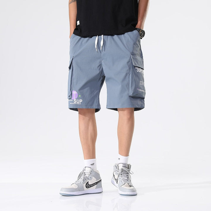 Un homme porte un short cargo bleu ample avec inscriptions. Il porte des baskets montantes blanches et un tee-shirt noir.