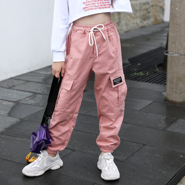 Une enfant pose en pantalon cargo taille haute resserré aux chevilles rose dans une rue et top manches longues blanc. Elle a des baskets blanches et un sac bandoulière violet