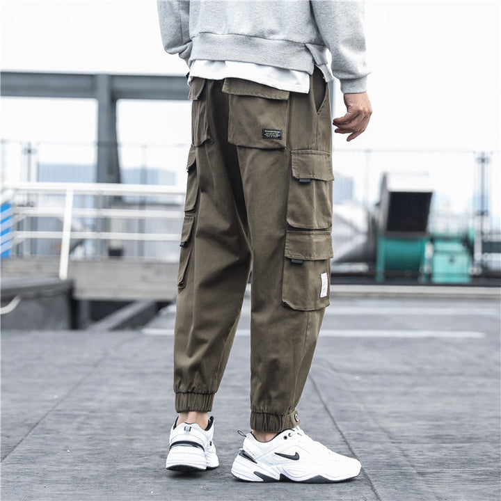 Un homme porte un pantalon cargo ample multipoches resserré aux chevilles de couleur taupe. Ce pantalon en coton 100% offre confort et durabilité. Disponible en différentes tailles, du S au 5XL.