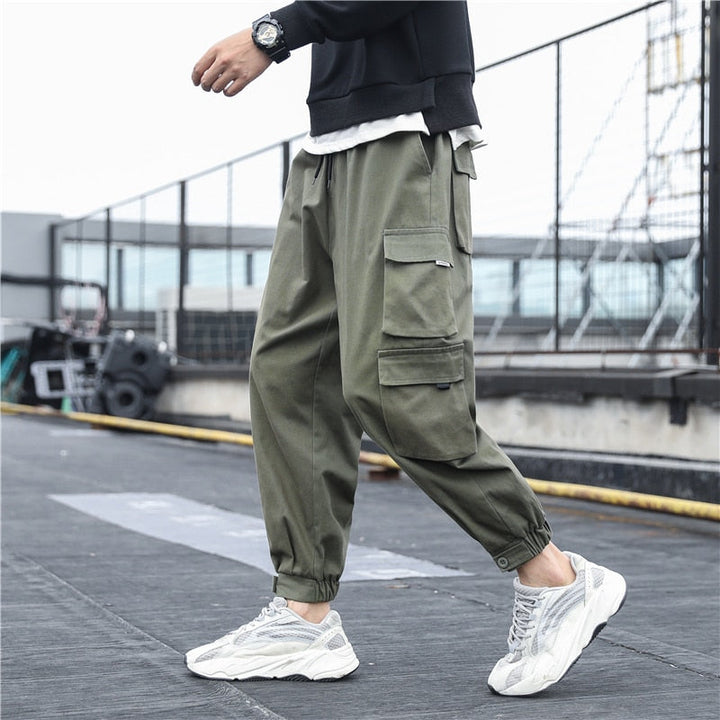 Un homme porte un pantalon cargo ample multipoches resserré aux chevilles de couleur kaki. Le pantalon est fabriqué en coton 100% pour un confort maximal et une durabilité garantie. Disponible en différentes tailles du S au 5XL.