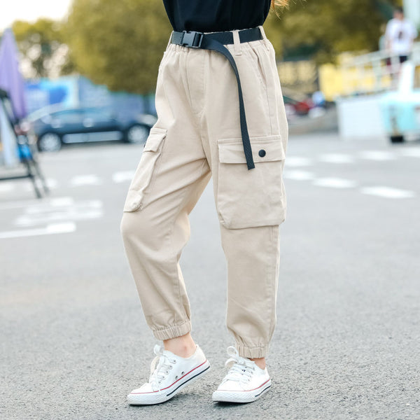 Une fille pose dans la rue. Elle porte un haut noir, des baskets basses blanches et un pantalon cargo jogger beige avec ceinture à clip noire.