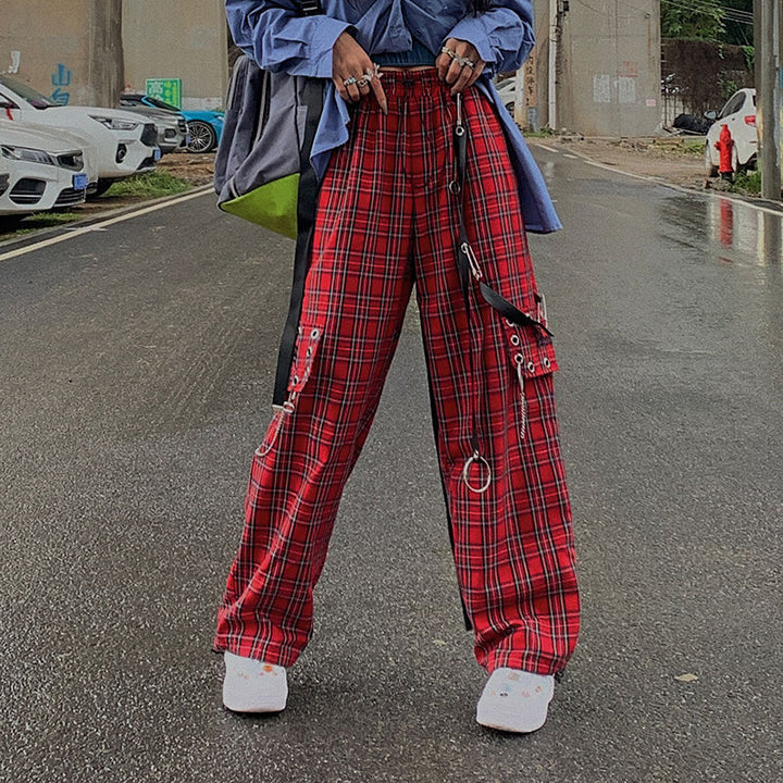 Une femme pose dans la rue avec une chemise en jean oversize, un sac à dos gris et vert, des baskets blanches et un pantalon cargo rouge à carreaux. Le pantalon est taille haute, évasé et il est orné de lanières en similicuir cuir avec épingles, oeillets et chaînes. Il donne un look très punk à la tenue.