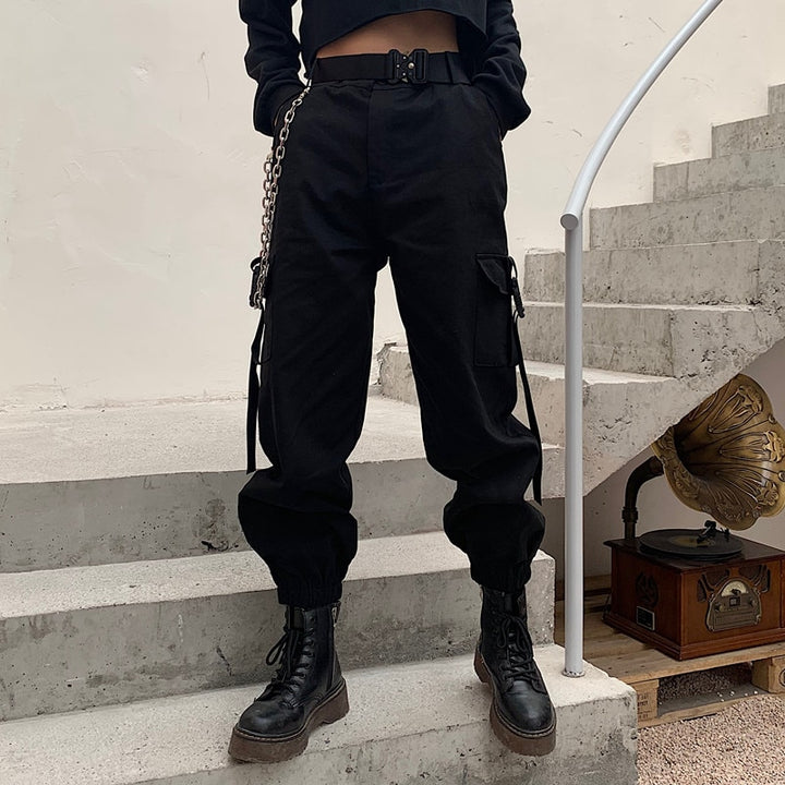 Une personne portant un pantalon cargo noir avec des poches à clip aux chevilles - Femme. Design urbain distinctif avec des poches latérales fermées par des clips en plastique. Disponible du S au 3XL. Coupe droite et taille haute mettant en valeur la silhouette.