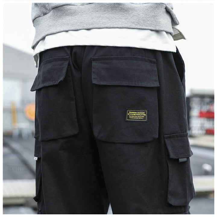 Un homme porte un pantalon cargo noir ample avec des poches multiples, resserré aux chevilles. Confortable et durable, ce pantalon en coton 100% est disponible dans une large gamme de tailles. Idéal pour un look tendance et pratique.