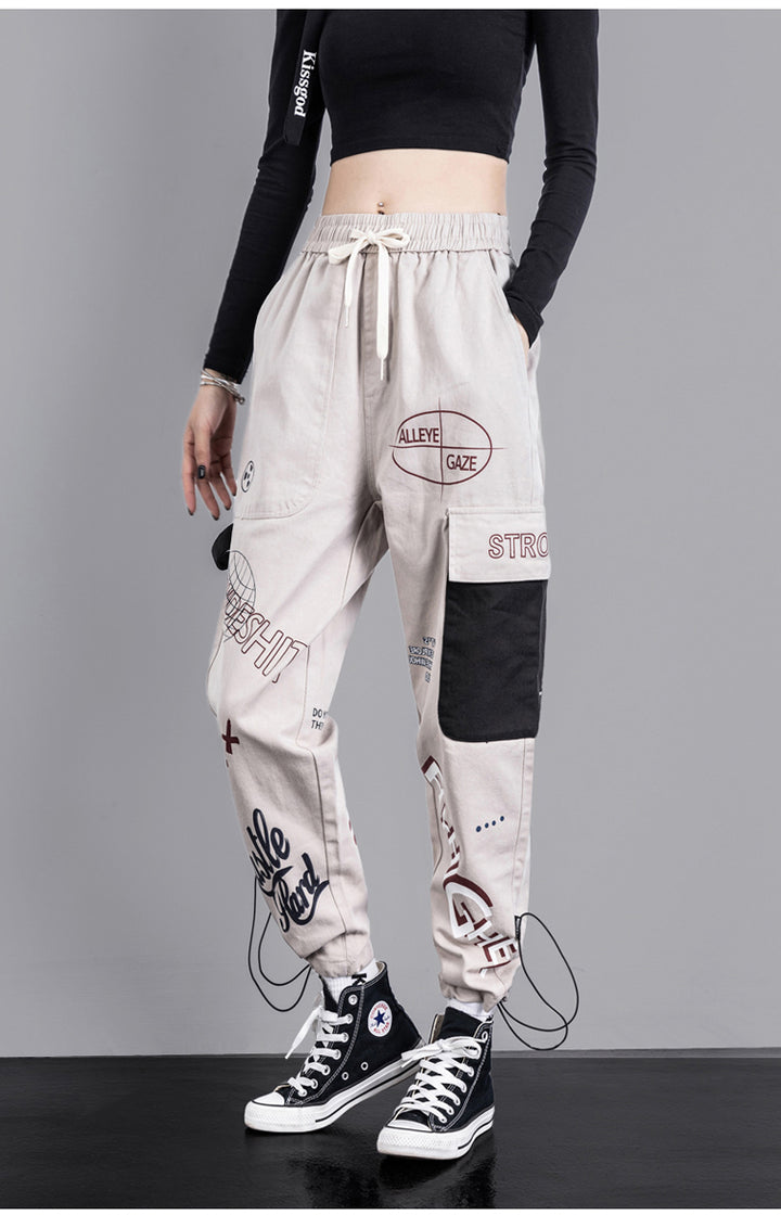 Une personne portant un pantalon cargo jogger gris clair à motifs noirs et roses. Le pantalon présente une coupe jogger taille haute avec une poche latérale et une lanière noires pour une allure urbaine. Disponible du S au 3XL, en coton pour un confort optimal.