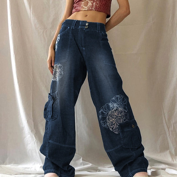 Une femme pose devant un tenture blanche. Elle porte un crop top rose et or avec un jean baggy taille basse avec un motif rosaces blanches. Le jean est bleu. Elle porte des baskets roses au pied. 