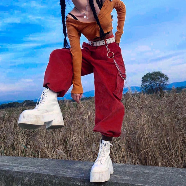 Une femme pose devant un champ de blé. Elle porte un gilet court orange, de grosses chaussures blanches ainsi qu'un pantalon cargo baggy en velours côtelé rouge avec chaînes sur le côté. Elle a les cheveux tressés.