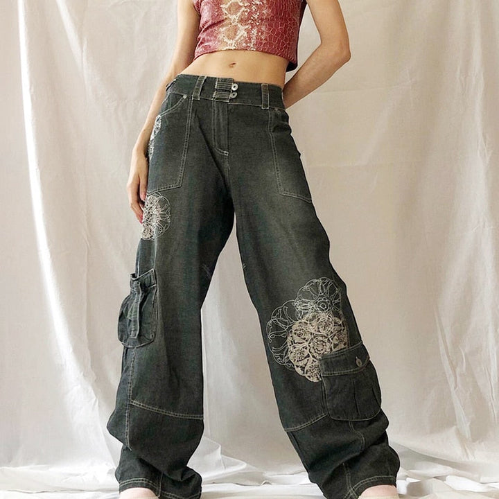 Une femme pose devant un tenture blanche. Elle porte un crop top rose et or avec un jean baggy taille basse avec un motif rosaces blanches. Le jean est gris. Elle porte des baskets roses au pied.