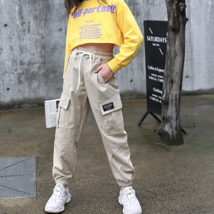 Une femme portant un pantalon cargo beige avec une coupe droite et des chevilles resserrées. Le pantalon est de taille haute avec des poches cargo fonctionnelles. Disponible en tailles 3 ans (110 cm) à 12 ans (160 cm). Parfait pour introduire le style streetwear dans la garde-robe de votre fille.