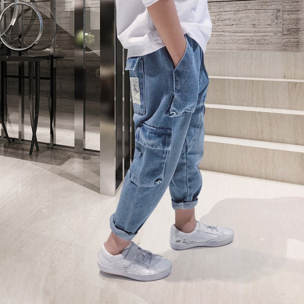 Un enfant pose devant des escaliers en marbre. Il porte un tee-shirt blanc, des baskets blanches ainsi qu'un pantalon cargo jean bleu. 