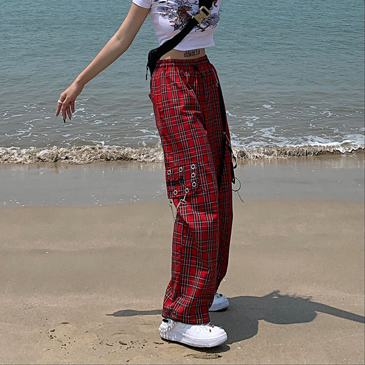 Femme en pantalon cargo à carreaux et chaînes sur la plage. Look rock avec coupe évasée et jambe large. Épingles et détails métalliques. Disponible du S au L.