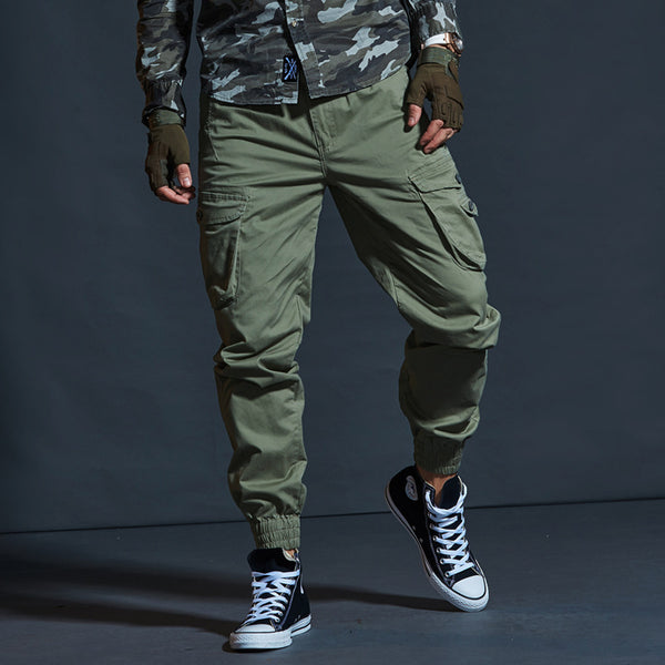 Un homme porte un pantalon cargo technique kaki, des converses noires, une chemise camo kaki et des mitaines kaki.