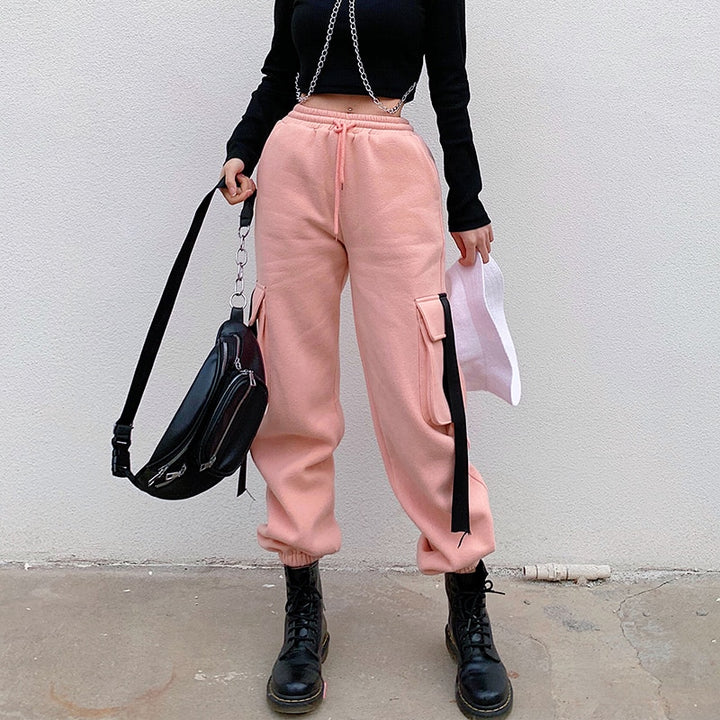 Une femme portant un pantalon cargo jogger rose avec des lanières noires sur les poches latérales. Taille haute, coupe jogger et élastique à la taille pour un confort et un style urbain. Disponible du S au L.