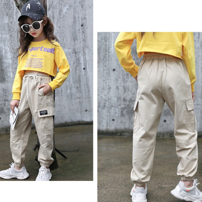 Une fille porte un pantalon cargo beige coupe droite avec des chevilles resserrées. Le pantalon est de style streetwear et dispose de poches cargo fonctionnelles. Taille haute avec élastique à la taille pour un ajustement parfait. Disponible en tailles 3 ans à 12 ans.