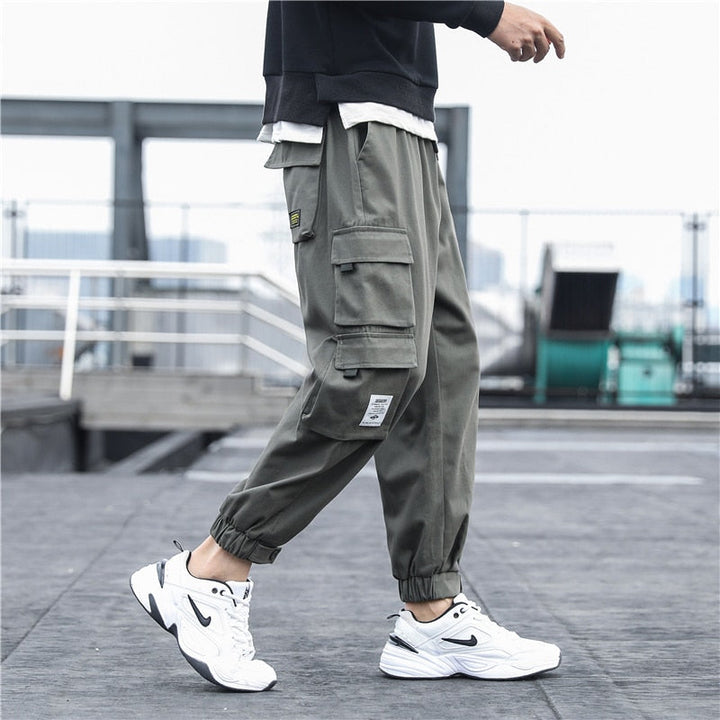 Un homme porte un pantalon cargo ample multipoches resserré aux chevilles gris. Le pantalon est en coton 100% pour un confort maximal et une durabilité garantie. Disponible en tailles S à 5XL.