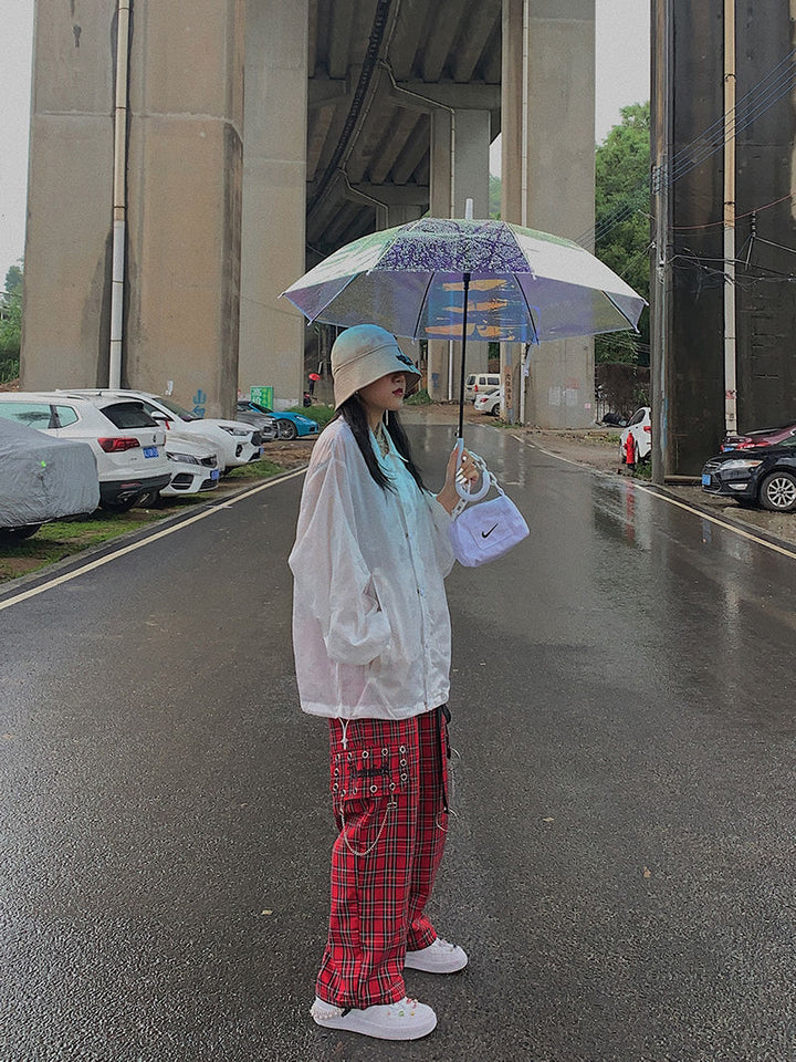 Une femme tenant un parapluie, portant un pantalon cargo à carreaux et chaînes rouge. Le pantalon a une coupe évasée et une jambe large, avec des détails rock tels que des épingles et des lanières en similicuir. Disponible du S au L.