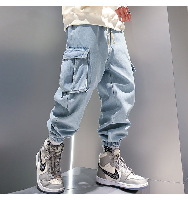 Un homme porte un jean cargo baggy resserré aux chevilles et des sneakers. Le jean en denim bleu offre un look streetwear affirmé et un maximum de confort. Disponible du M au 5XL.