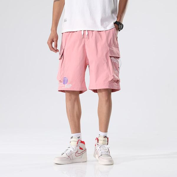 Un homme porte un short cargo rose ample avec inscriptions. Il porte des baskets montantes blanches et un tee-shirt blanc.