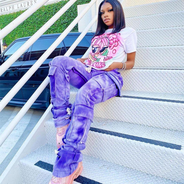 Une femme noire pose dans un escalier métallique. Elle porte un tee-shirt blanc à motif rose, des baskets roses ainsi qu'un pantalon cargo jogger tie and dye violet.