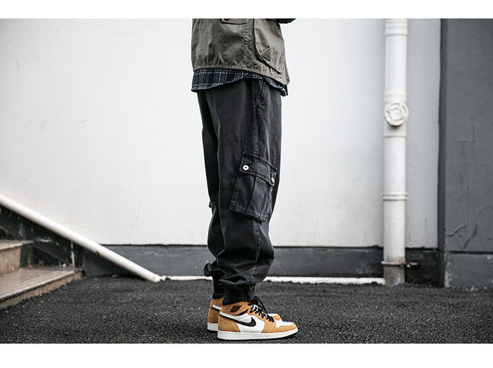 Un homme porte un jean cargo jogger coupe large noir avec des chaussures orange et blanches. Le jean est en toile denim, confortable et durable. Les poches latérales inclinées ajoutent une touche unique à ce style cargo. Tailles disponibles du S au 2XL.