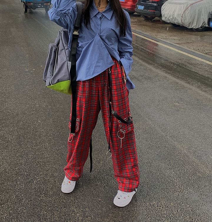 Une femme debout dans la rue portant un Pantalon cargo à carreaux et chaînes rouge. Le pantalon taille haute affiche une coupe évasée et une jambe large, orné d'une chaîne métallique, d’œillets en métal et de lanières en similicuir. Des épingles sur les côtés ajoutent une touche décalée. Disponible du S au L.