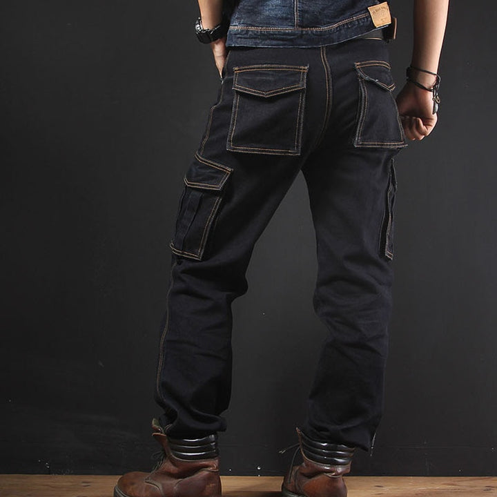 Un homme pose de dos dans une pièce aux murs noirs et parquet au sol. Il porte une veste en jean, des boots marrons et un jean cargo coupe droite noir.