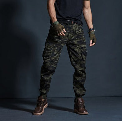 Un homme porte un pantalon cargo technique kaki camo, des boots marrons, un tee-shirt noir et des mitaines kakis.