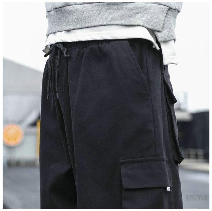 Un homme porte un pantalon cargo noir ample avec des poches multiples et resserré aux chevilles. Confortable et durable, il est fabriqué en coton 100%. Tailles disponibles du S au 5XL.