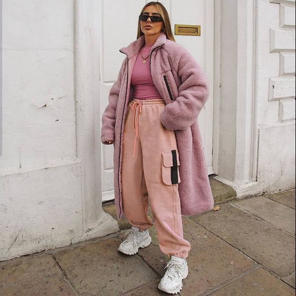 Une femme blonde avec des lunettes de soleil pose dans la rue. Elle porte un manteau rose, un tee-shirt rose, des baskets blanches et un pantalon cargo jogger rose avec des lanières noires sur les poches latérales. 