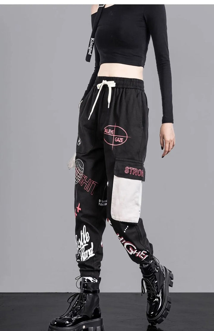 Une femme portant un pantalon cargo jogger noir à motifs blancs et roses, avec une poche latérale et une lanière gris clair. Taille haute et coupe ample resserrée aux chevilles pour un confort optimal. Disponible du S au 3XL. Pantalon cargo jogger à motif - Noir - Femme.